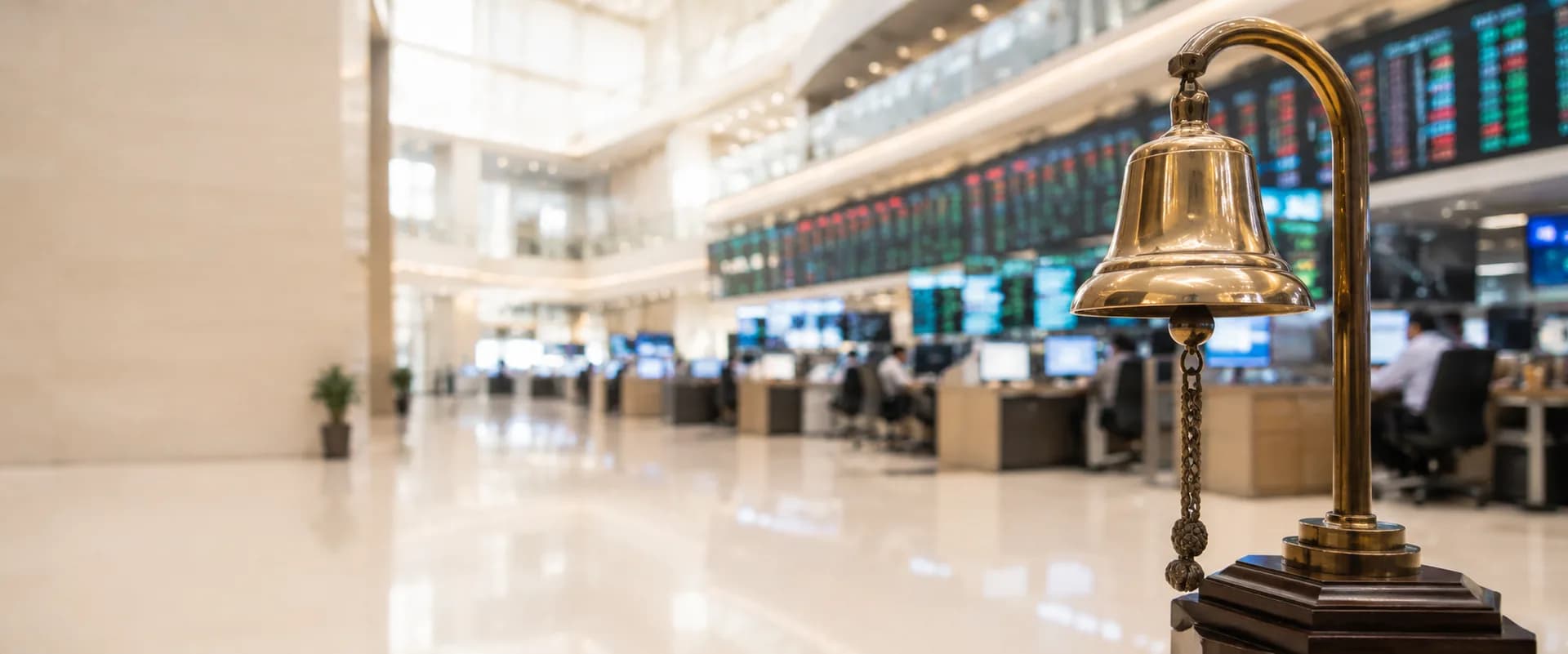 A brass listing ceremony bell in the foreground of a stock exchange hall, with trading screens behind it