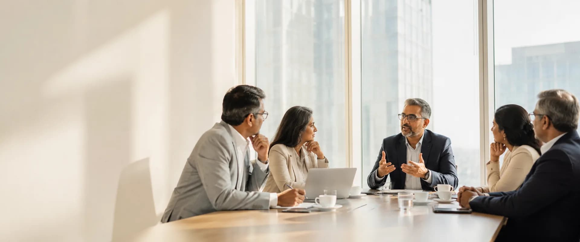 Five senior advisors in discussion around a boardroom table in a sunlit office overlooking the city